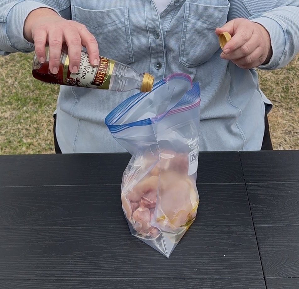 A person pours vinegar from a bottle into a resealable plastic bag containing raw chicken pieces, with the bag resting on a black table outdoors.
