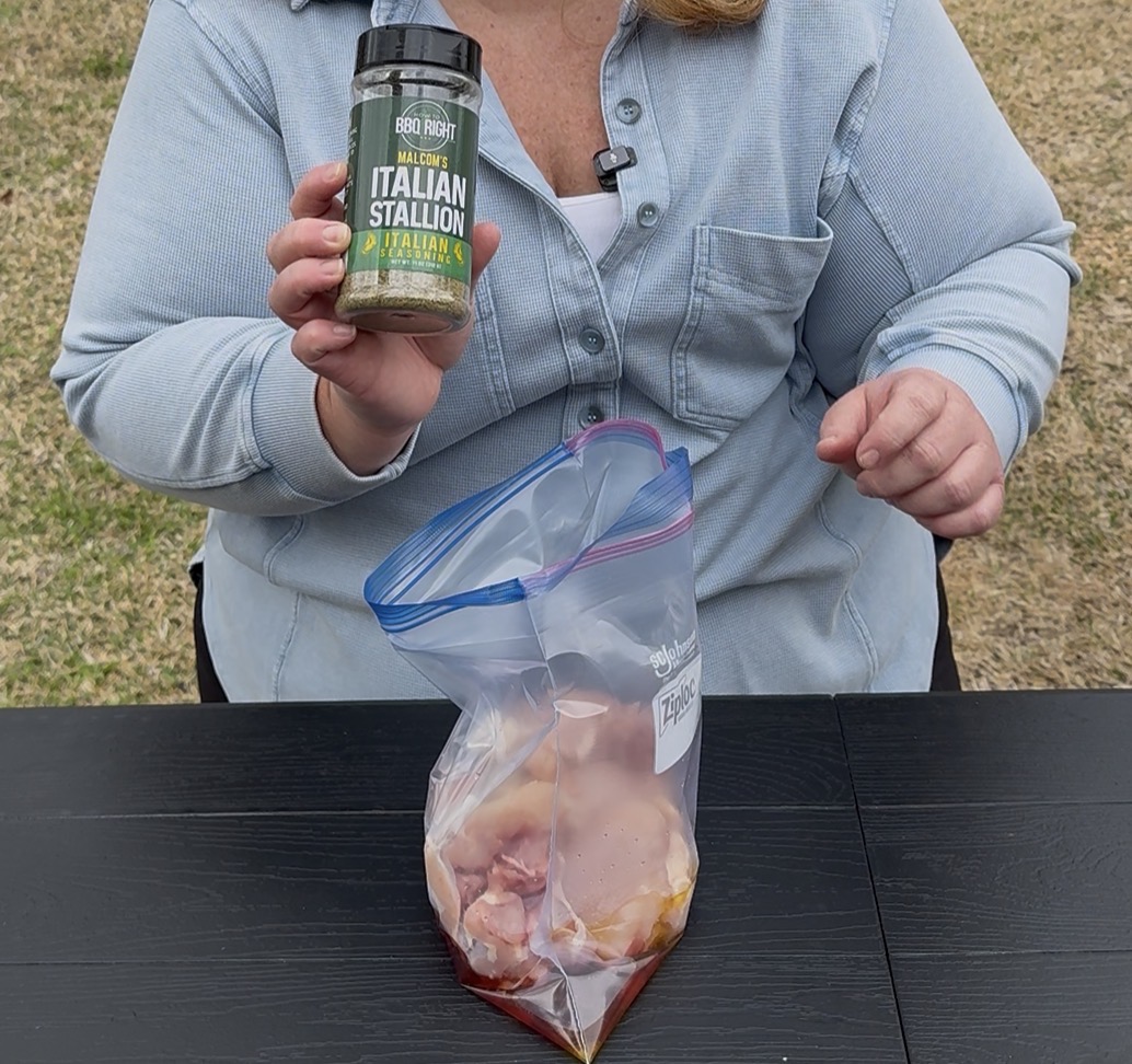 A person in a blue shirt holds a jar of BBQ Right Italian Stallion seasoning above a plastic bag containing raw chicken pieces on a black table outdoors.
