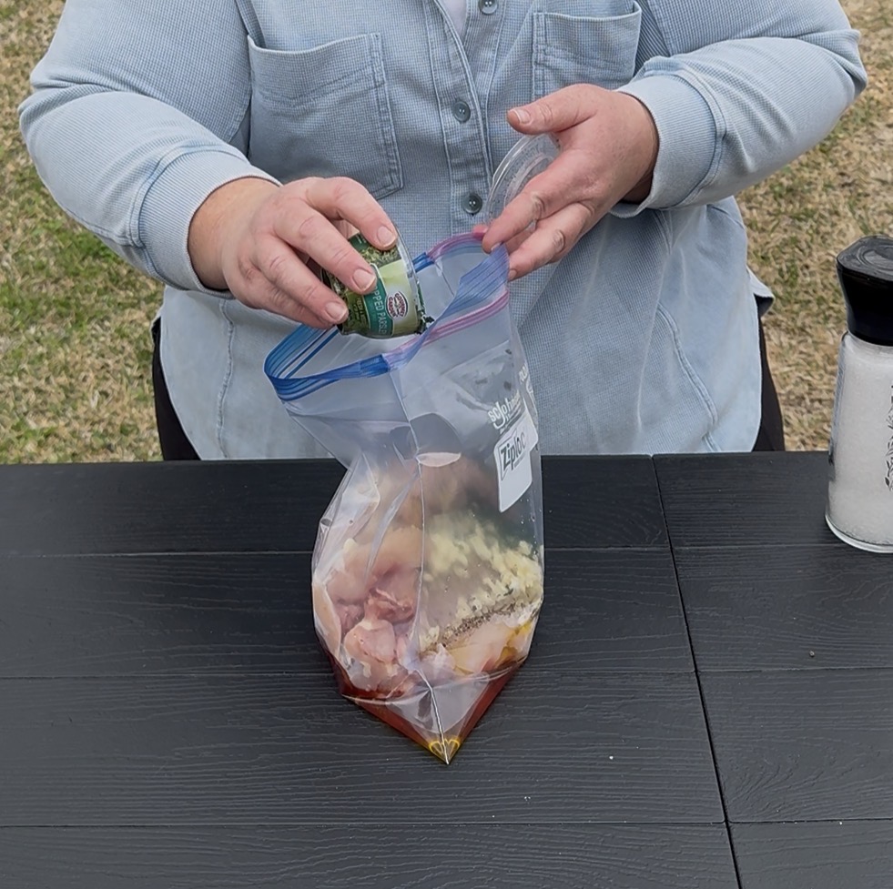 A person pours a can of soda into a resealable plastic bag containing raw chicken and marinade ingredients, on a black table outdoors. A container of salt is on the table nearby.