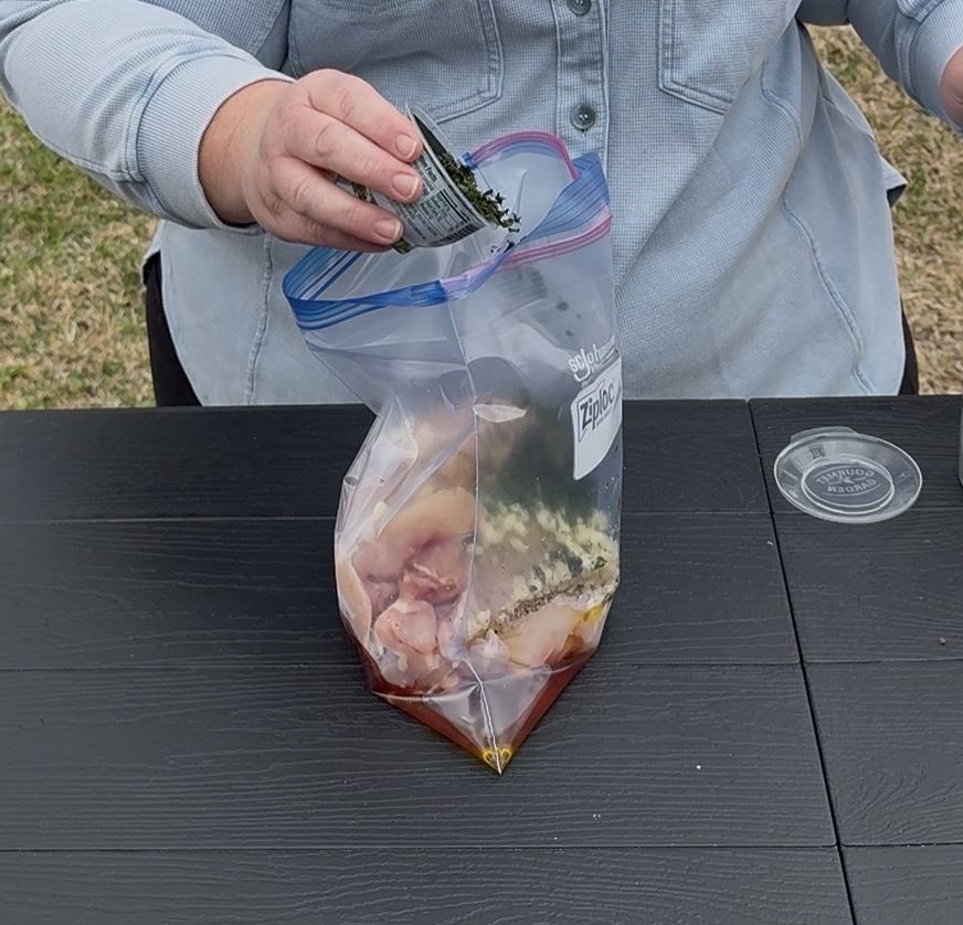 A person is pouring seasoning from a packet into a resealable plastic bag containing raw chicken and marinade ingredients on a black table outdoors.