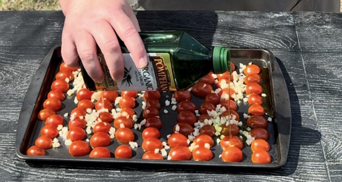 A hand pours olive oil from a bottle over a baking tray filled with halved cherry tomatoes and chopped garlic, arranged neatly on a dark surface.