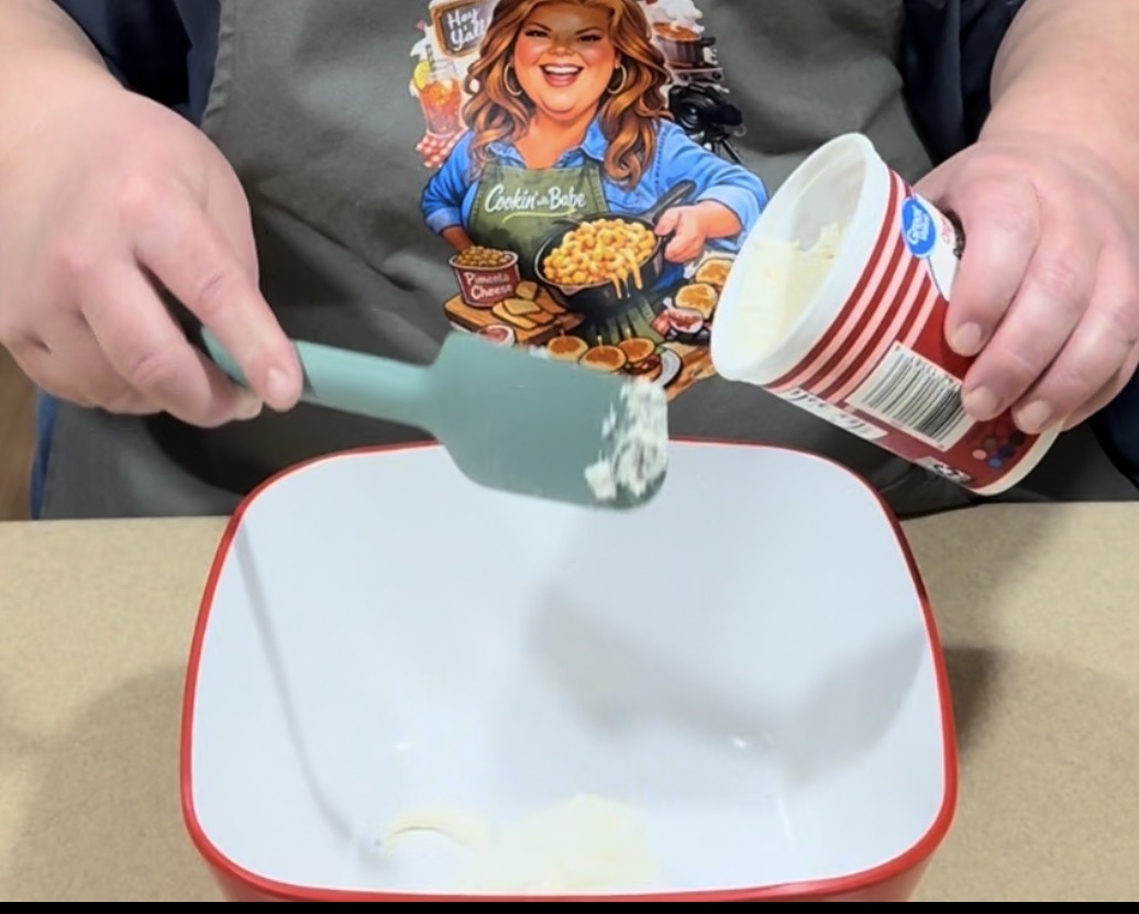 A person wearing an apron with a cartoon chef design uses a blue spatula to scoop white powder, likely flour, from a striped container into a square white bowl on a counter.