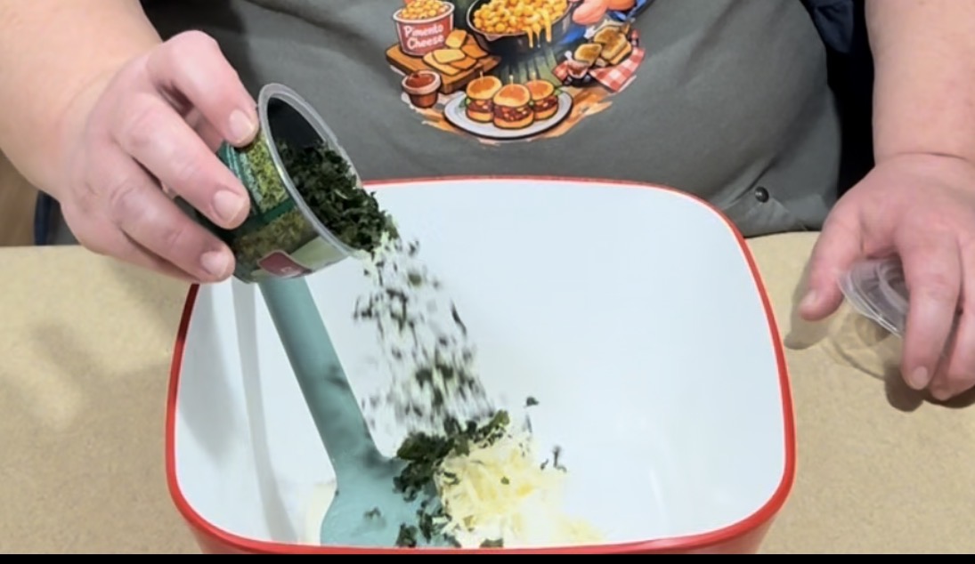 A person pours chopped spinach from a can into a white mixing bowl containing shredded cheese, using a teal spatula. The person is wearing a shirt with an illustrated food-themed design.