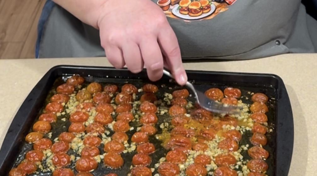 A person uses a fork to mash roasted cherry tomatoes and garlic on a baking tray.
