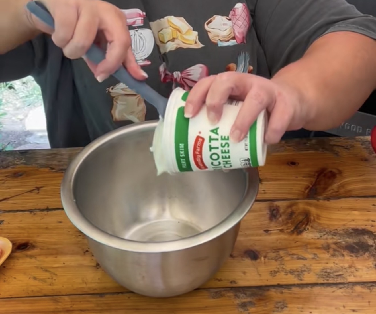 A person scoops cottage cheese from a container into a metal mixing bowl on a wooden table. The person is wearing a dark shirt with colorful kitchen utensil and ingredient graphics.