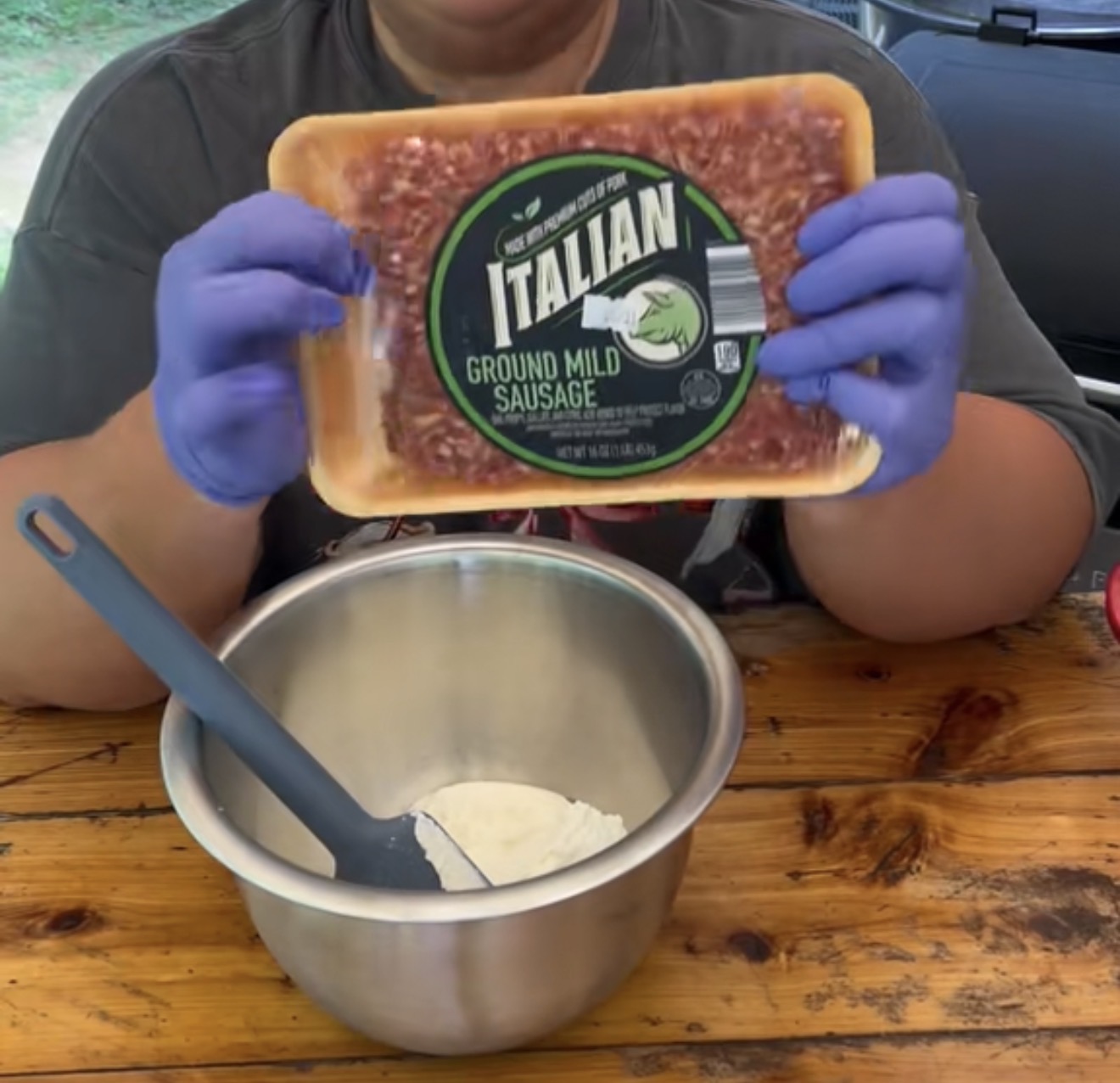 A person wearing blue gloves holds a package of Italian ground mild sausage above a metal mixing bowl with a black spatula, all placed on a wooden table.
