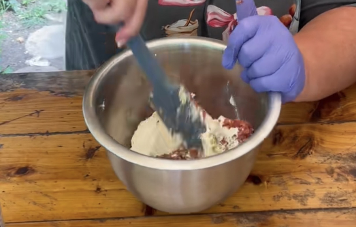 A person wearing a patterned shirt and a purple glove mixes ingredients, including flour, in a large metal bowl with a spatula on a wooden table.