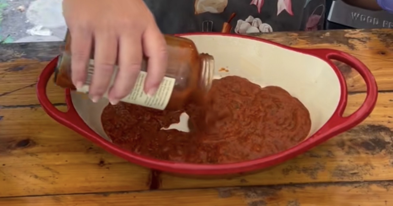 A hand pours tomato sauce from a jar into a red and white oval baking dish on a wooden surface.