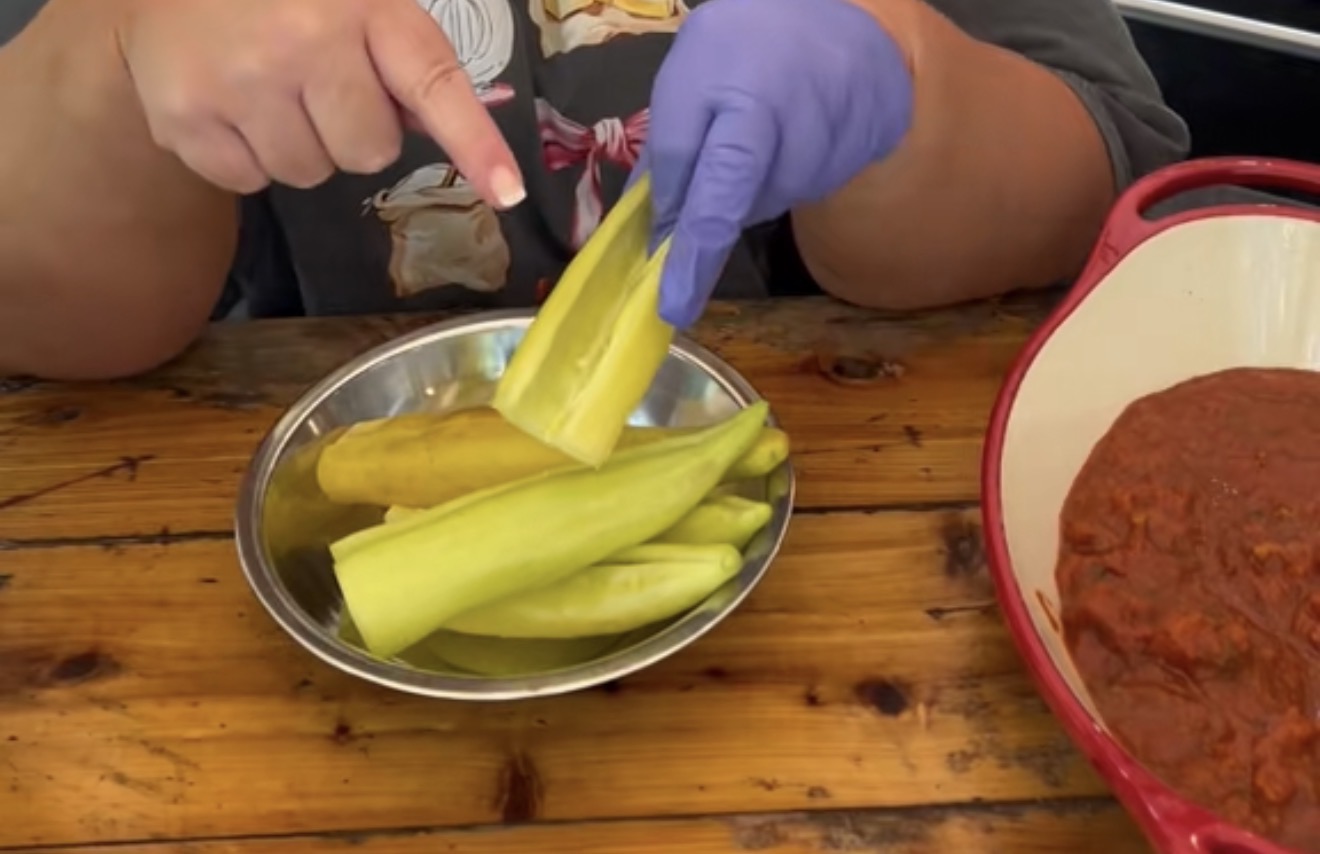 A person wearing a glove holds and points to a hollowed-out pale green pepper over a metal bowl filled with similar peppers; a red dish with a chunky red sauce sits nearby on a wooden table.