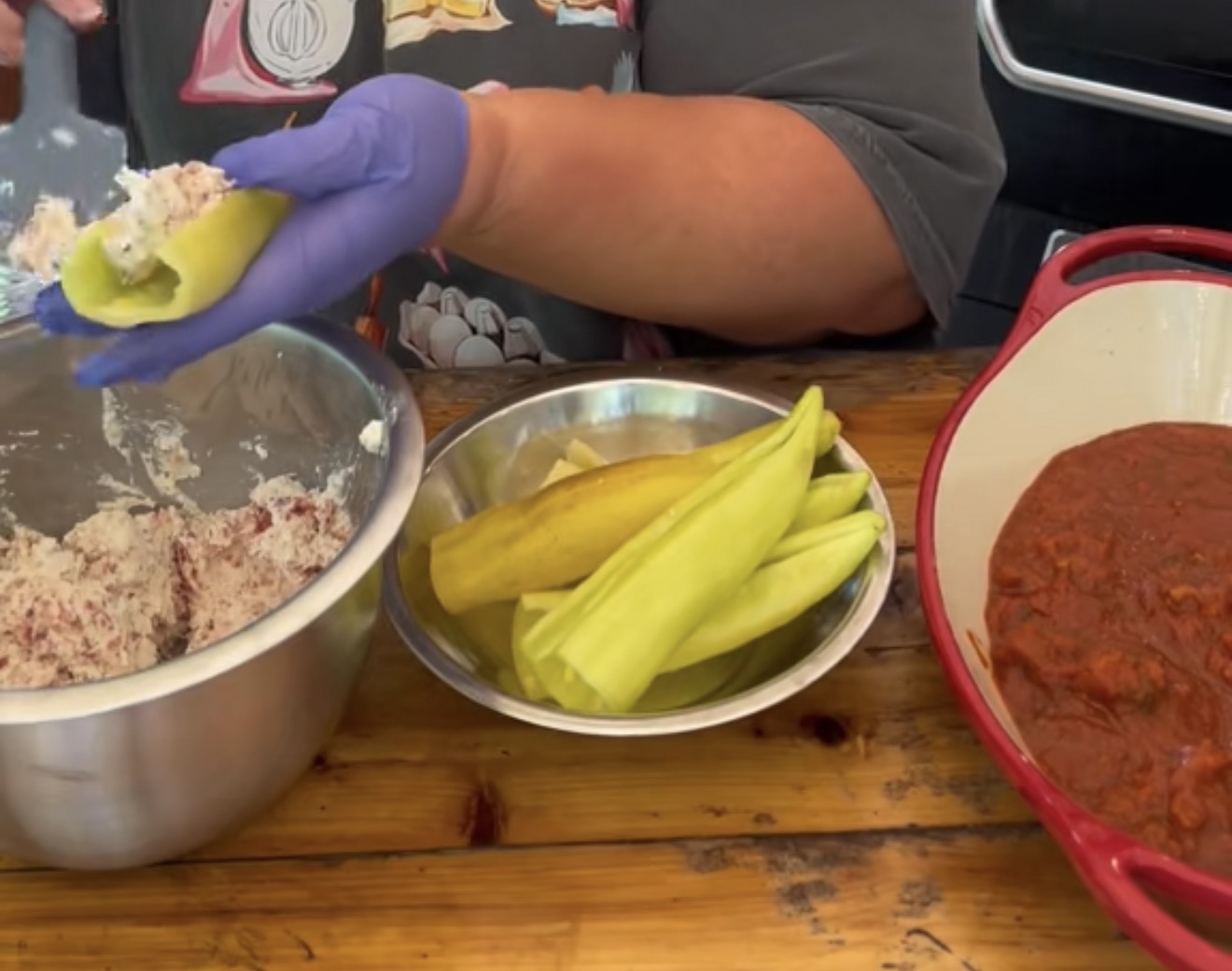 A person wearing gloves stuffs pale green peppers with a rice filling. The table holds a metal bowl of filling, a plate of peppers, and a red pot of tomato sauce.