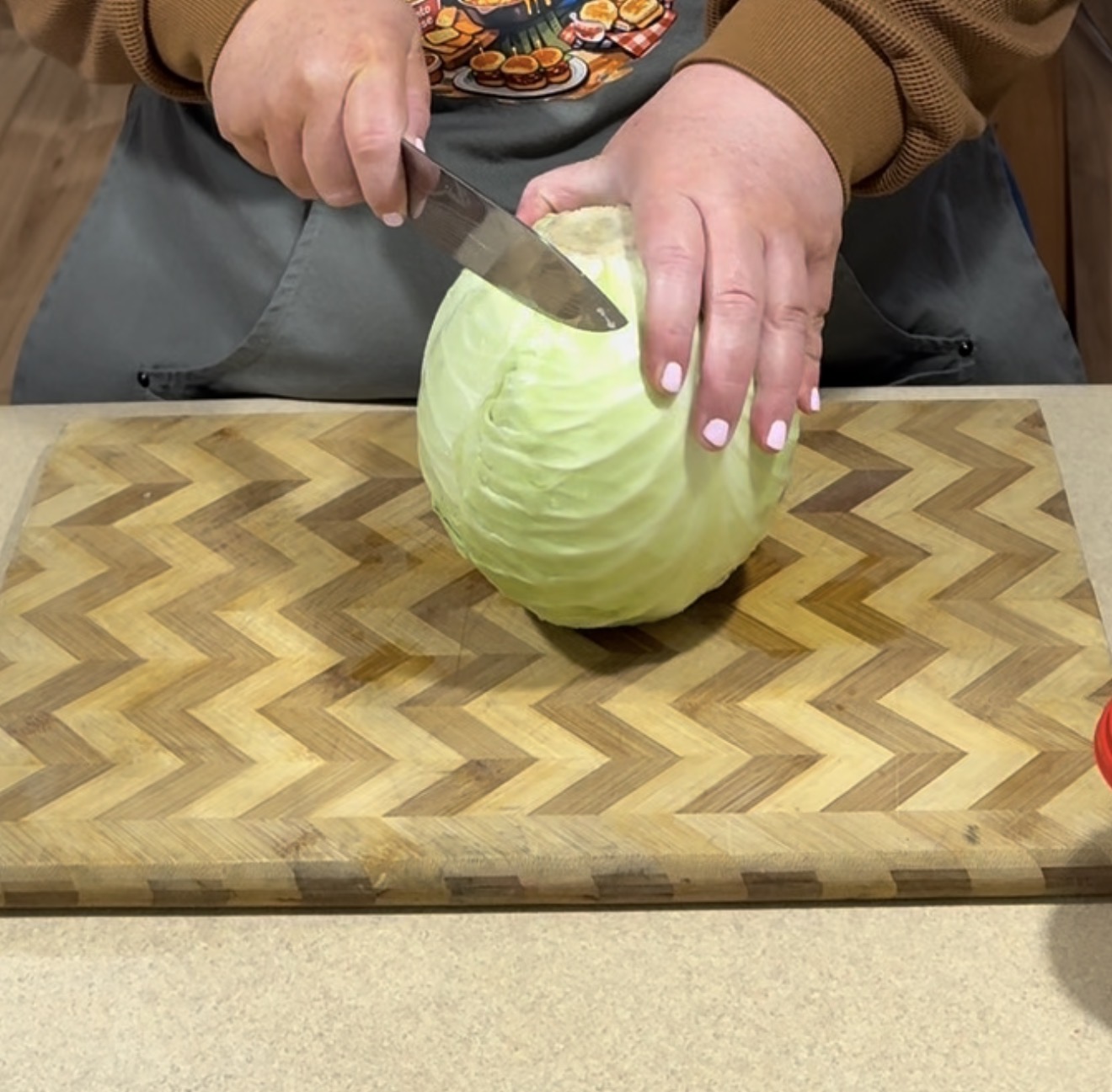 A person cutting a green cabbage on a zigzag-patterned wooden cutting board, holding the cabbage steady with one hand and slicing with a kitchen knife in the other hand.