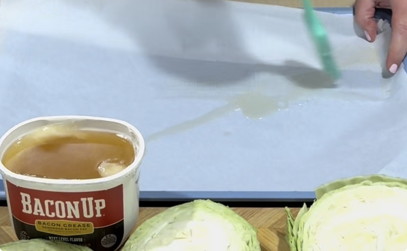 A person brushes a baking sheet lined with parchment paper using bacon grease from a Bacon Up container. Halved cabbage heads are visible on a cutting board nearby.