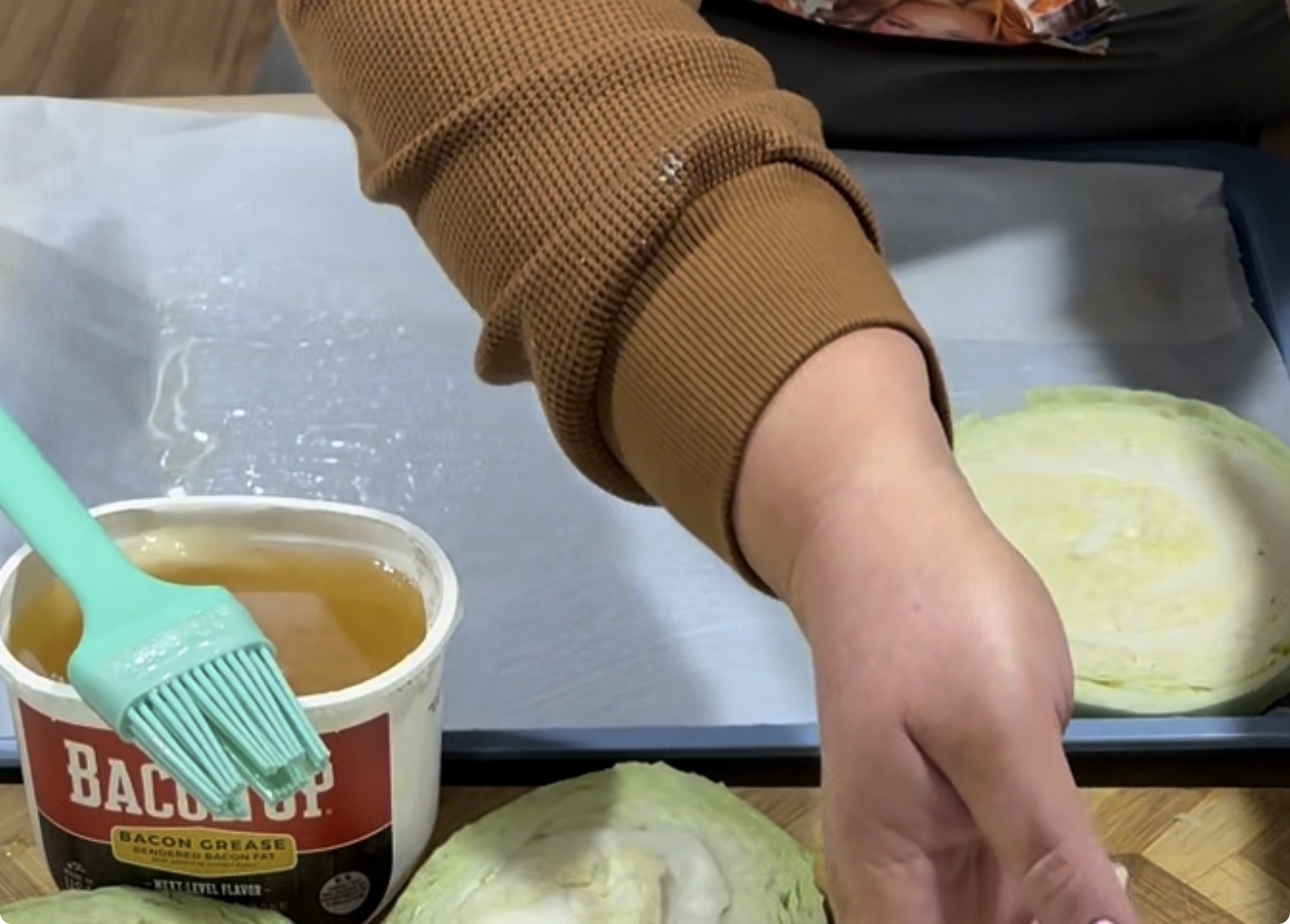 A person in a brown sweater brushes bacon grease onto a baking sheet lined with parchment paper. Slices of cabbage and a container labeled Bacon Grease with a green brush are on the table.