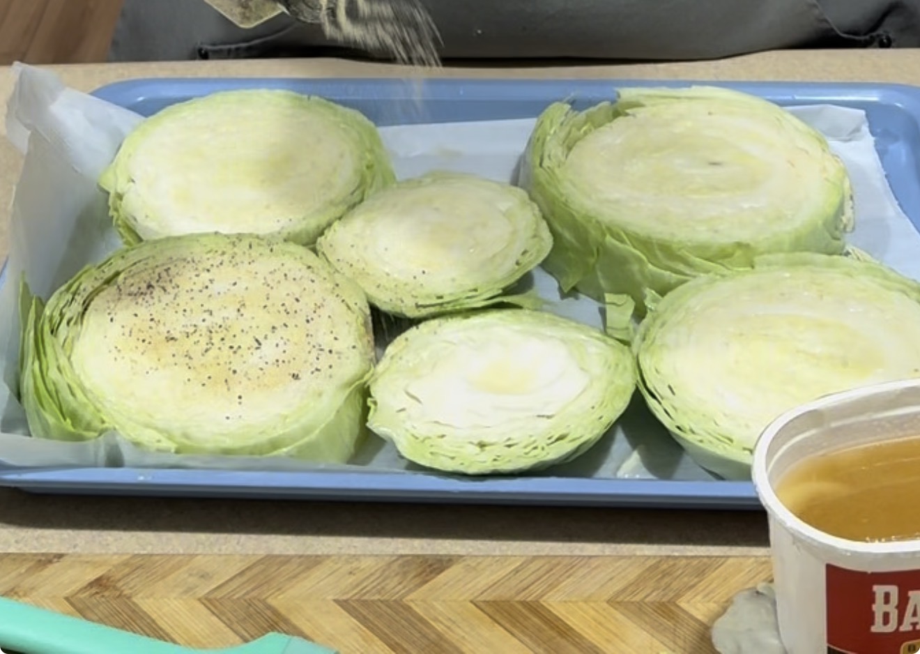 Sliced cabbage rounds on a blue baking tray, some sprinkled with black pepper. A parchment sheet lines the tray, and a cup with a brown liquid is visible in the foreground.