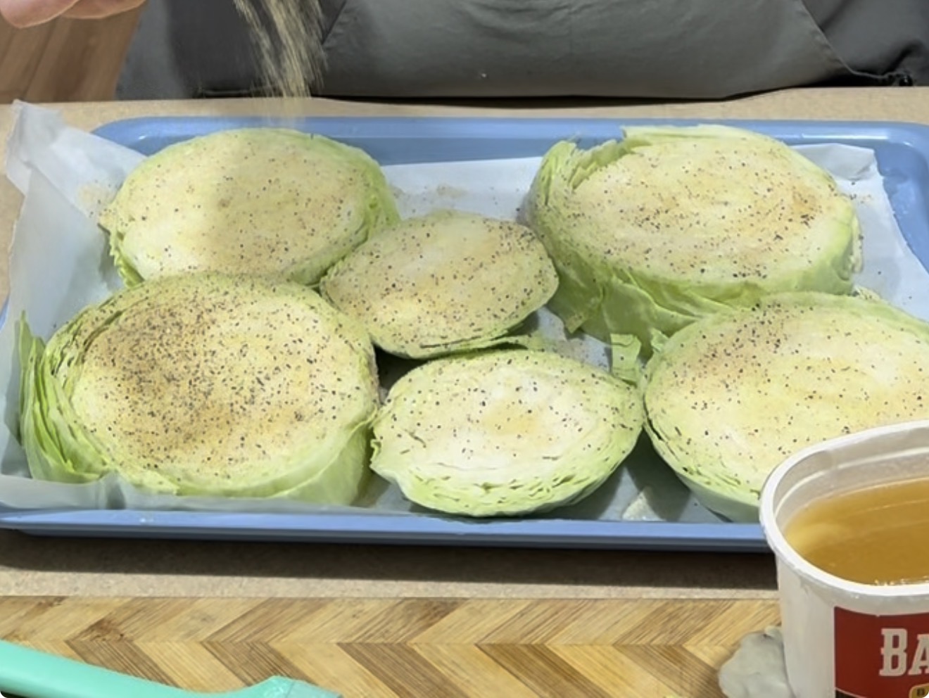 Sliced cabbage rounds on a baking tray are being sprinkled with seasoning. A container of broth and a spatula are visible nearby on a wooden cutting board.