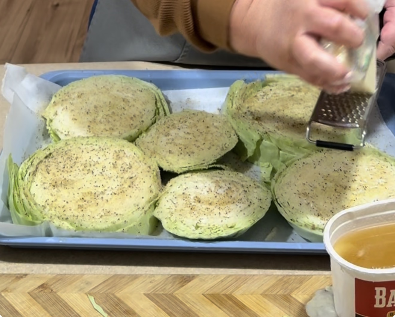A person grates cheese over seasoned cabbage slices arranged on a parchment-lined baking tray. A container with liquid and a wooden cutting board are nearby.
