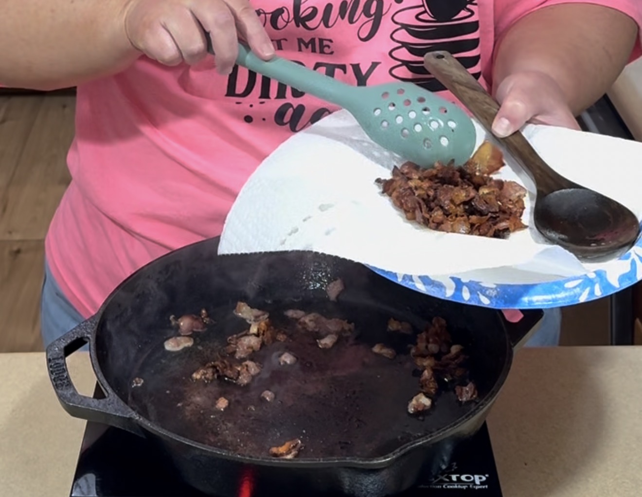 A person in a pink shirt uses a slotted spatula to transfer cooked bacon pieces from a cast iron skillet onto a paper towel-lined plate. A wooden spoon rests on the plate.