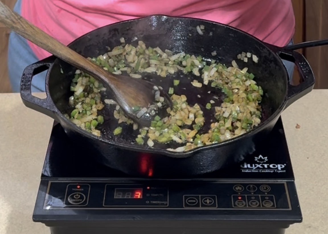 A person stirs chopped onions and green peppers in a cast iron skillet using a wooden spoon on an electric stovetop.