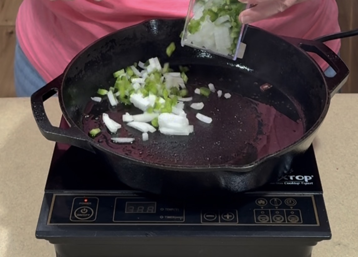 A person in a pink shirt adds chopped green bell pepper and onions from a small container into a black cast-iron skillet on an electric stovetop.