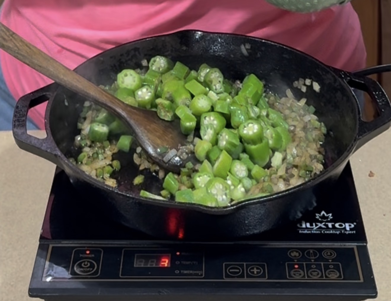 A cast iron skillet on an induction cooktop contains chopped okra and diced onions being sautéed with a wooden spoon. Steam rises from the vegetables, and a person in a pink shirt is visible behind the pan.