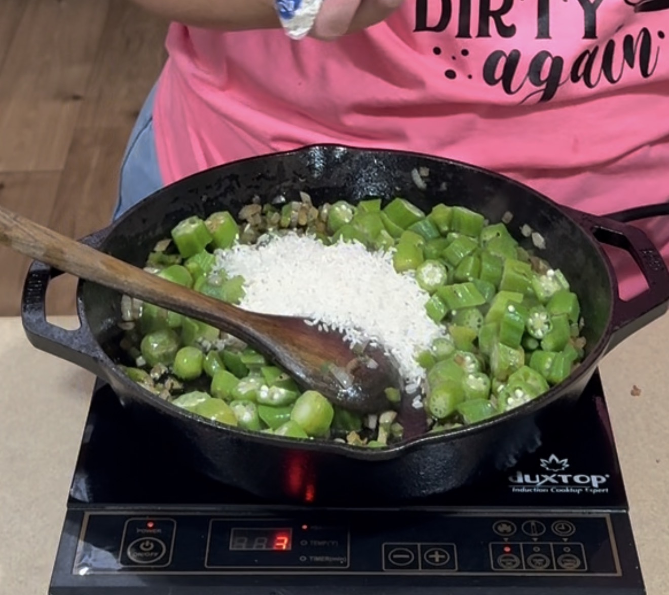 A person in a pink shirt stirs chopped okra, onions, and white rice in a cast iron skillet on an induction cooktop using a wooden spoon.