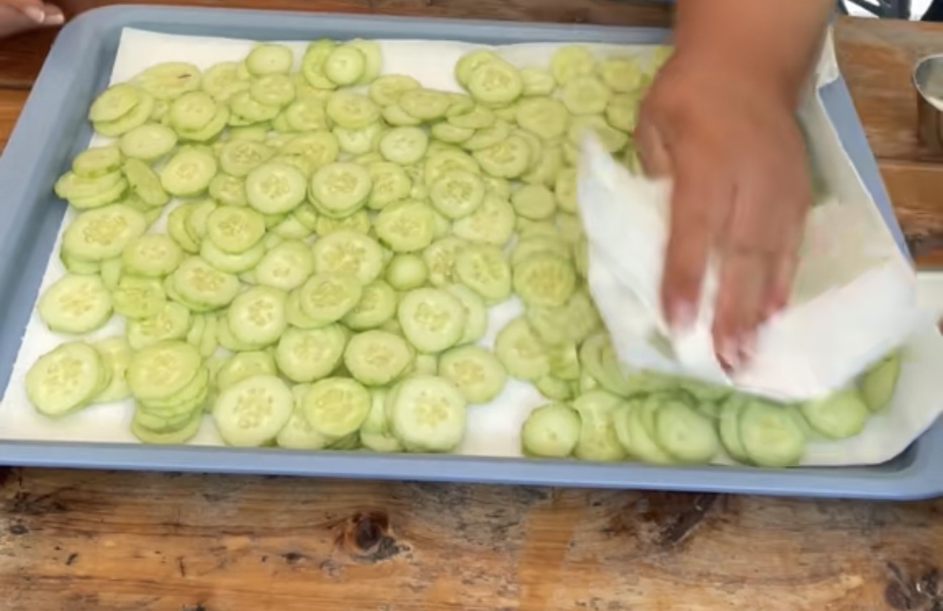 A hand uses a paper towel to blot moisture from sliced cucumbers arranged on a tray lined with paper towels, set on a wooden surface.