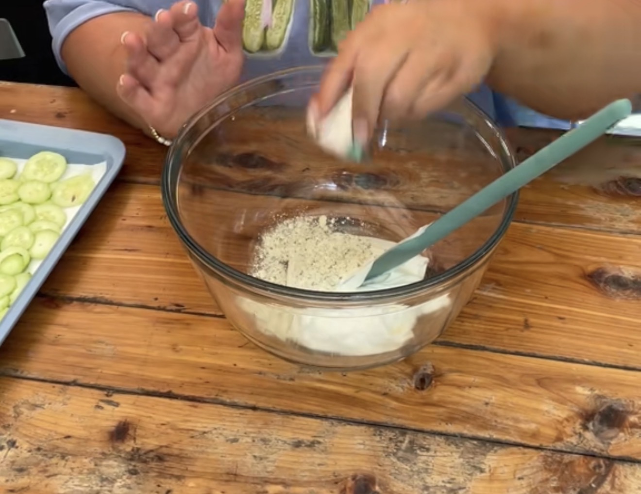 A person adds seasoning to a glass bowl with a creamy mixture, using their left hand while holding a spatula in their right hand; sliced cucumbers are on a tray nearby on a rustic wooden table.