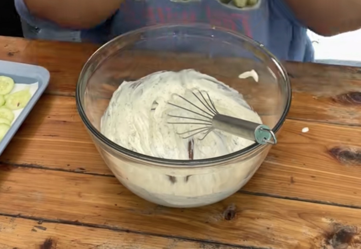 A glass bowl filled with a creamy white mixture and a whisk sits on a wooden table, with a tray of sliced cucumbers partially visible to the side.