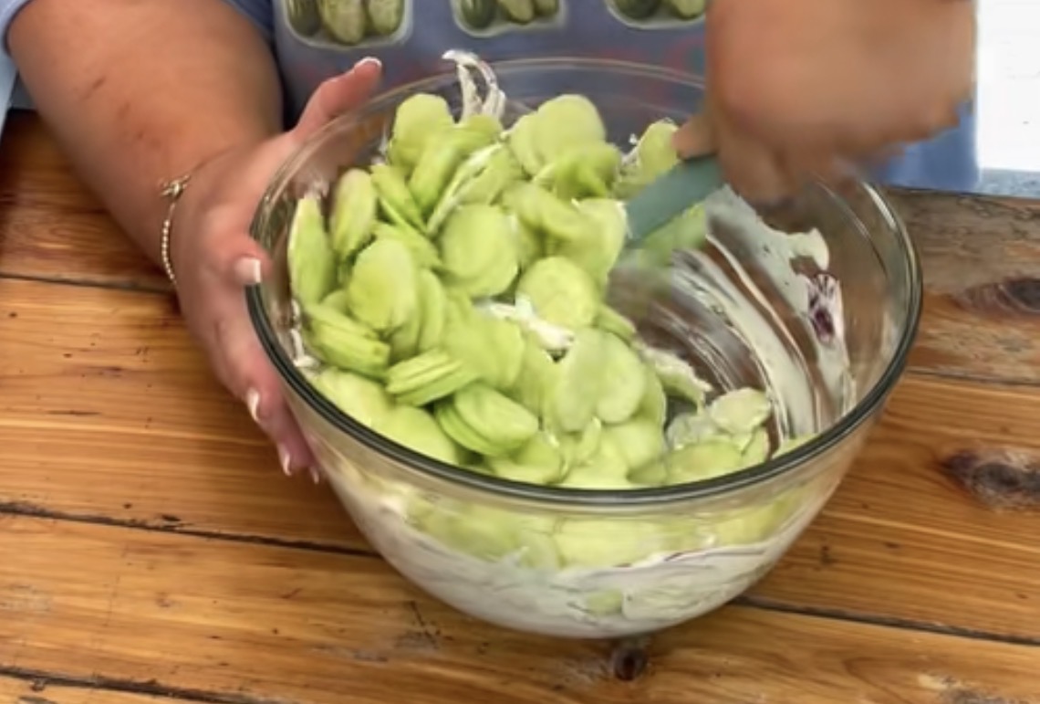 A person stirs sliced cucumbers with a creamy white dressing in a large glass bowl on a wooden table.