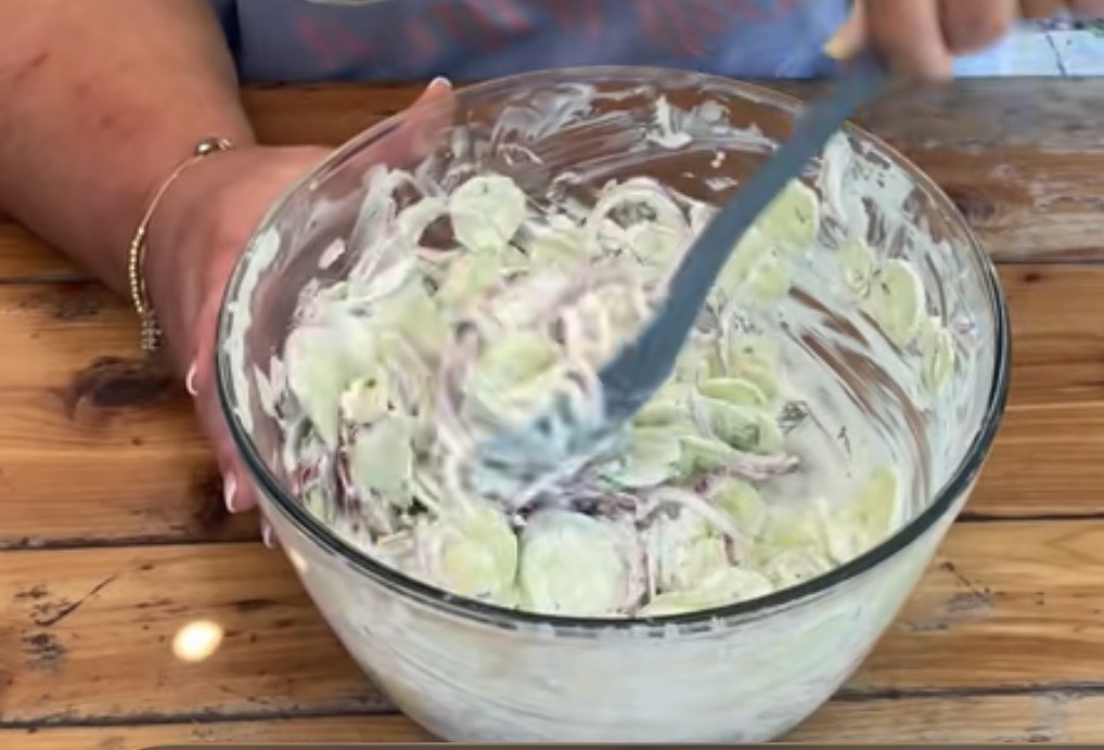 A person is stirring a bowl of creamy cucumber salad with a spoon on a wooden table. The salad contains sliced cucumbers and onions coated in a white dressing.