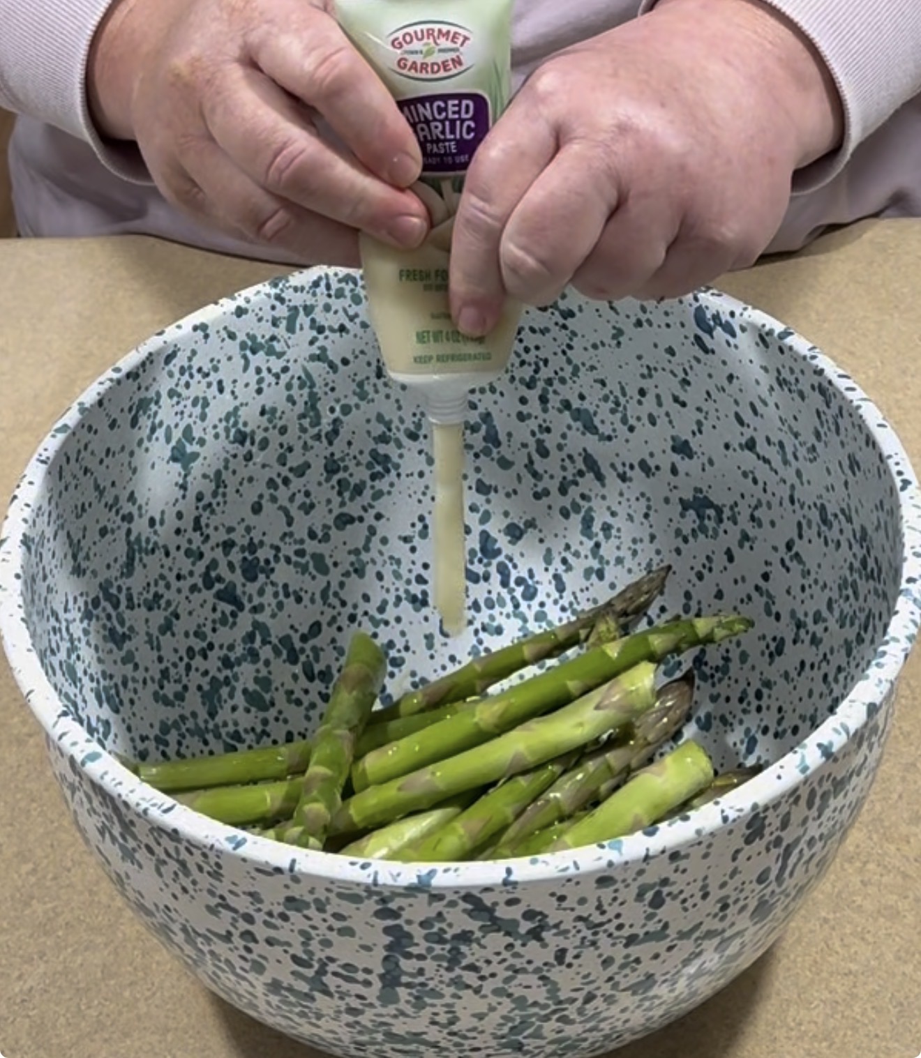 A person squeezes a tube of minced garlic over fresh asparagus spears in a speckled blue and white mixing bowl.