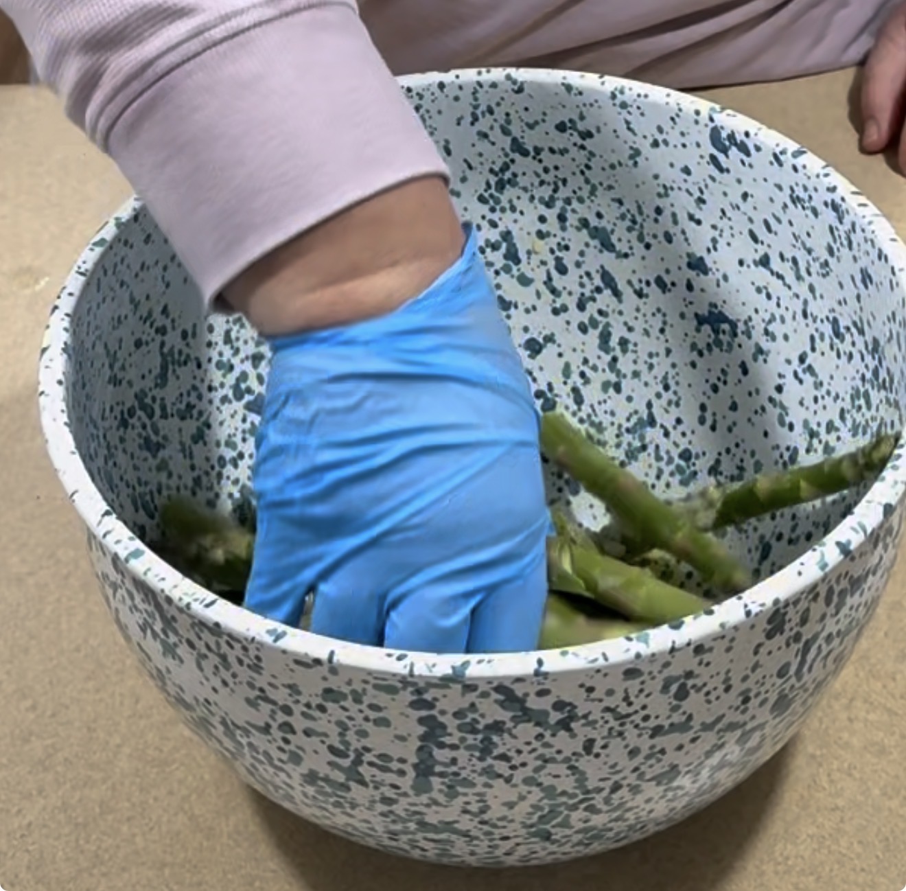 A person wearing a blue glove mixes or handles green vegetables in a speckled white and blue mixing bowl placed on a countertop.
