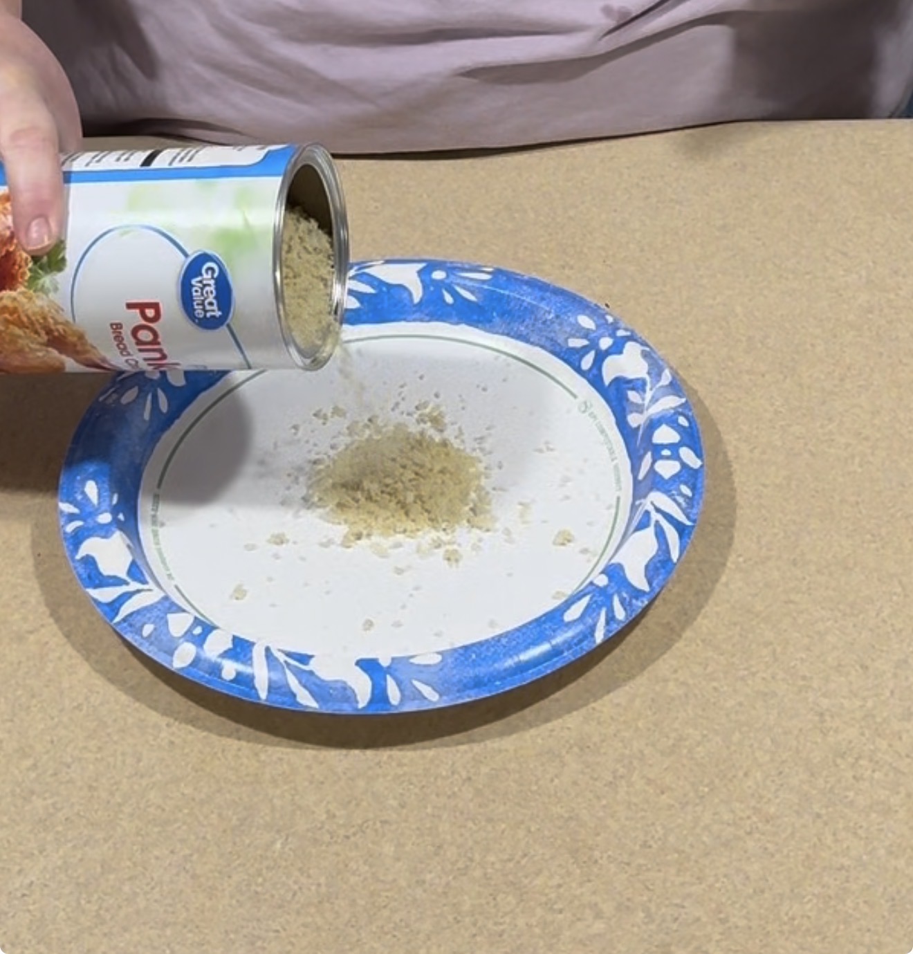 A person pours panko bread crumbs from a can onto a blue and white paper plate on a tan surface.