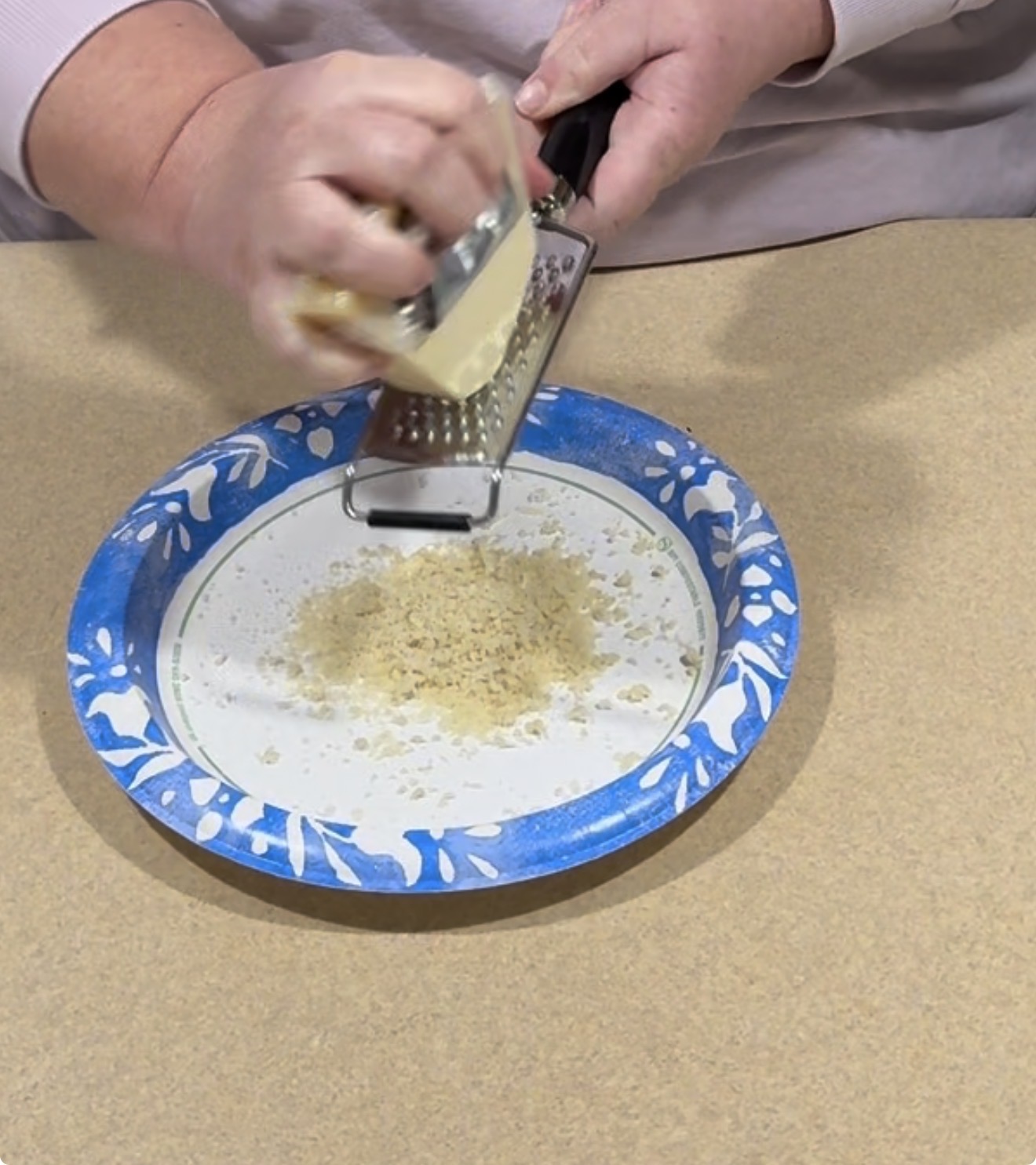 A person grates cheese over a decorative paper plate, with grated cheese piling up on the plate. Only their hands and part of their arms are visible.