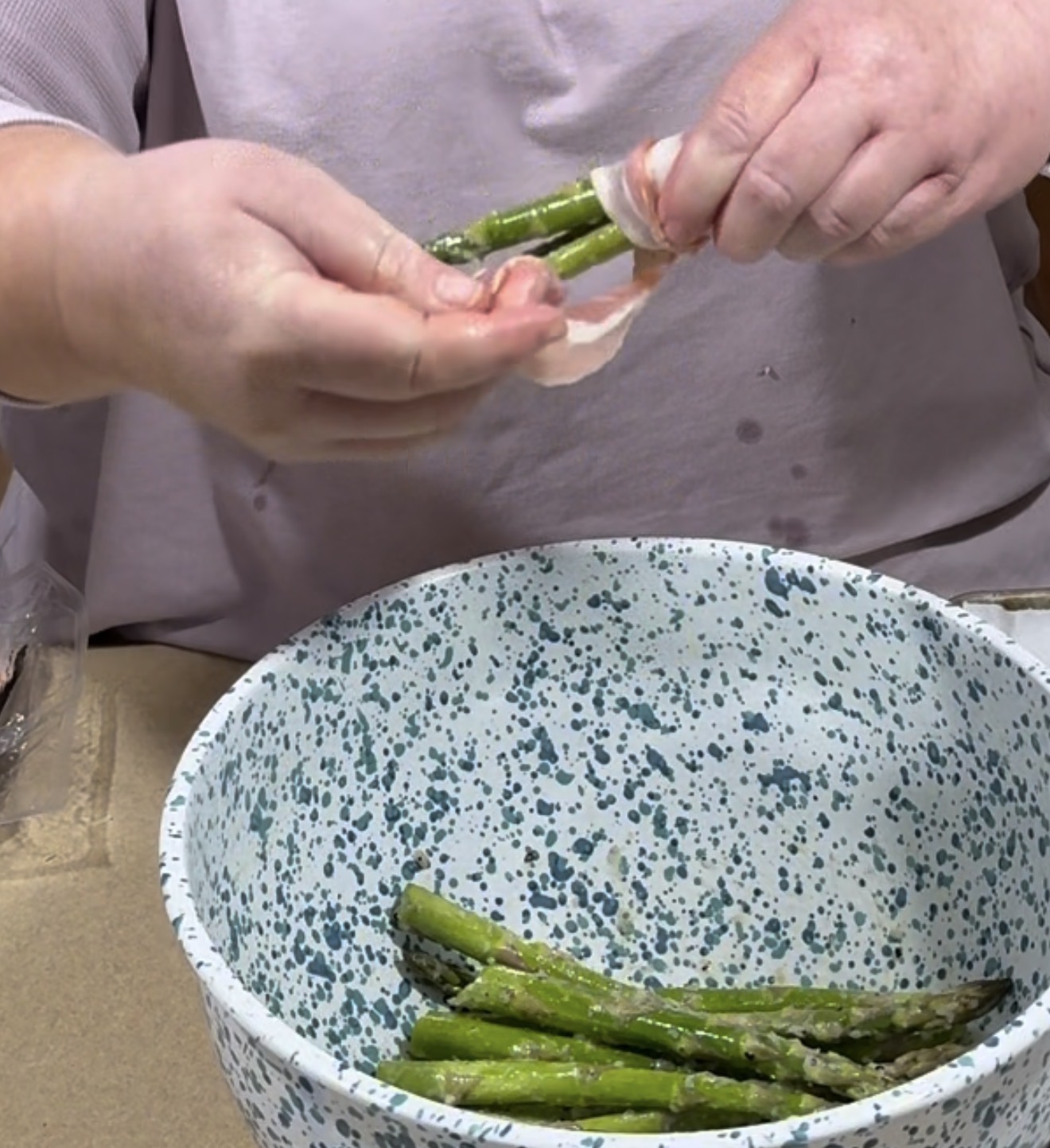 A person wraps raw bacon around asparagus spears over a blue and white speckled bowl containing more seasoned asparagus.