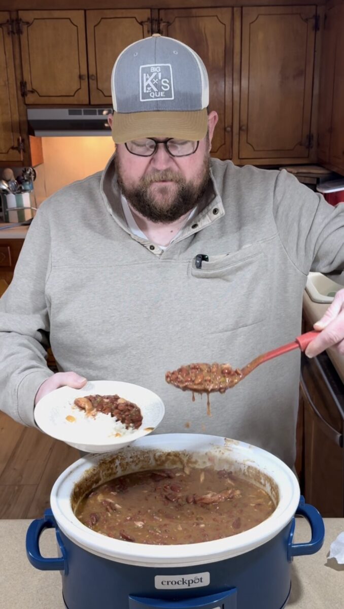 A man wearing glasses and a cap serves a scoop of thick chili from a crockpot into a white bowl in a kitchen with wooden cabinets in the background.