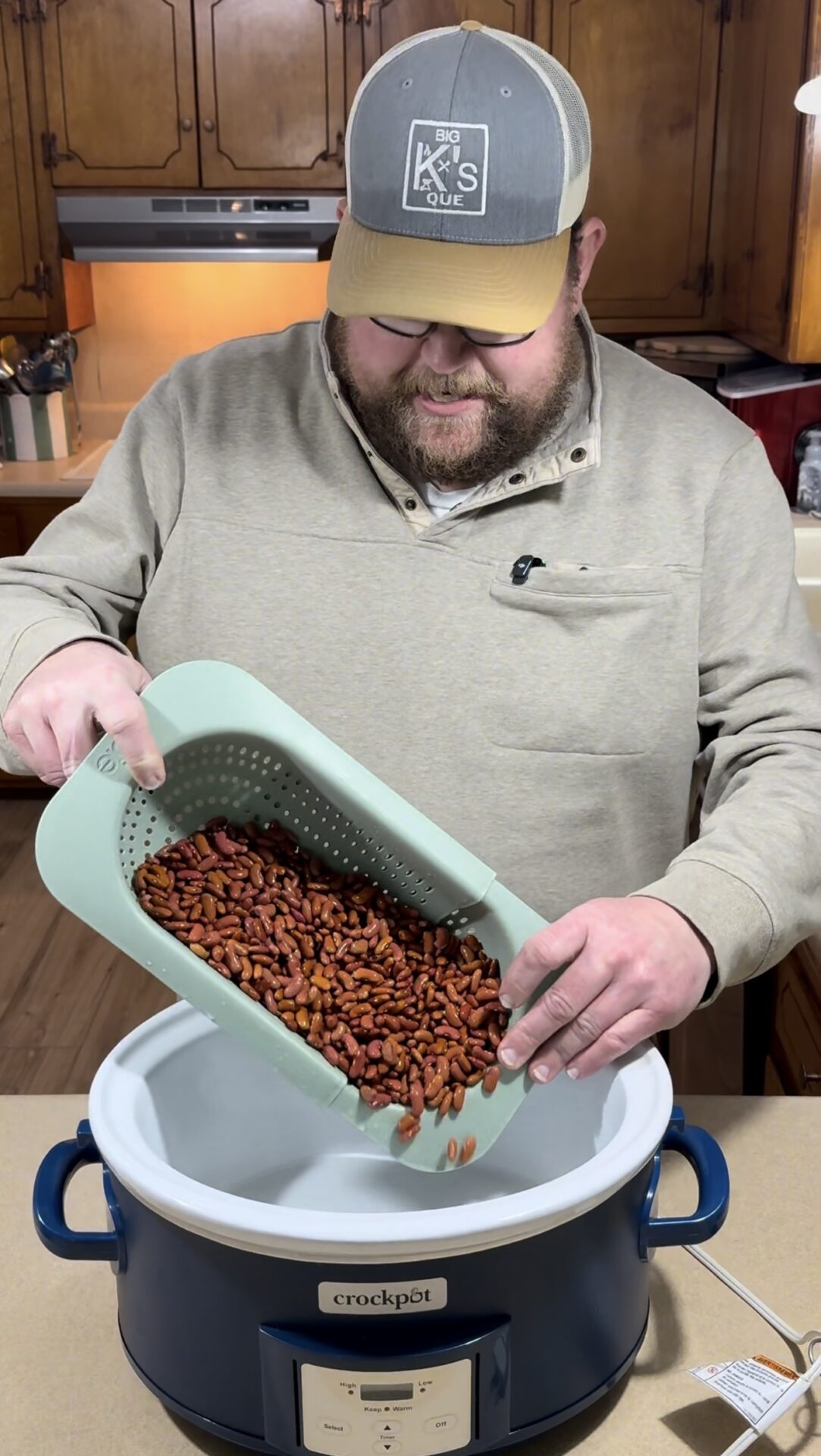 A man in a beige sweatshirt and cap pours rinsed red beans from a green colander into a blue and white Crockpot slow cooker in a kitchen with wooden cabinets.