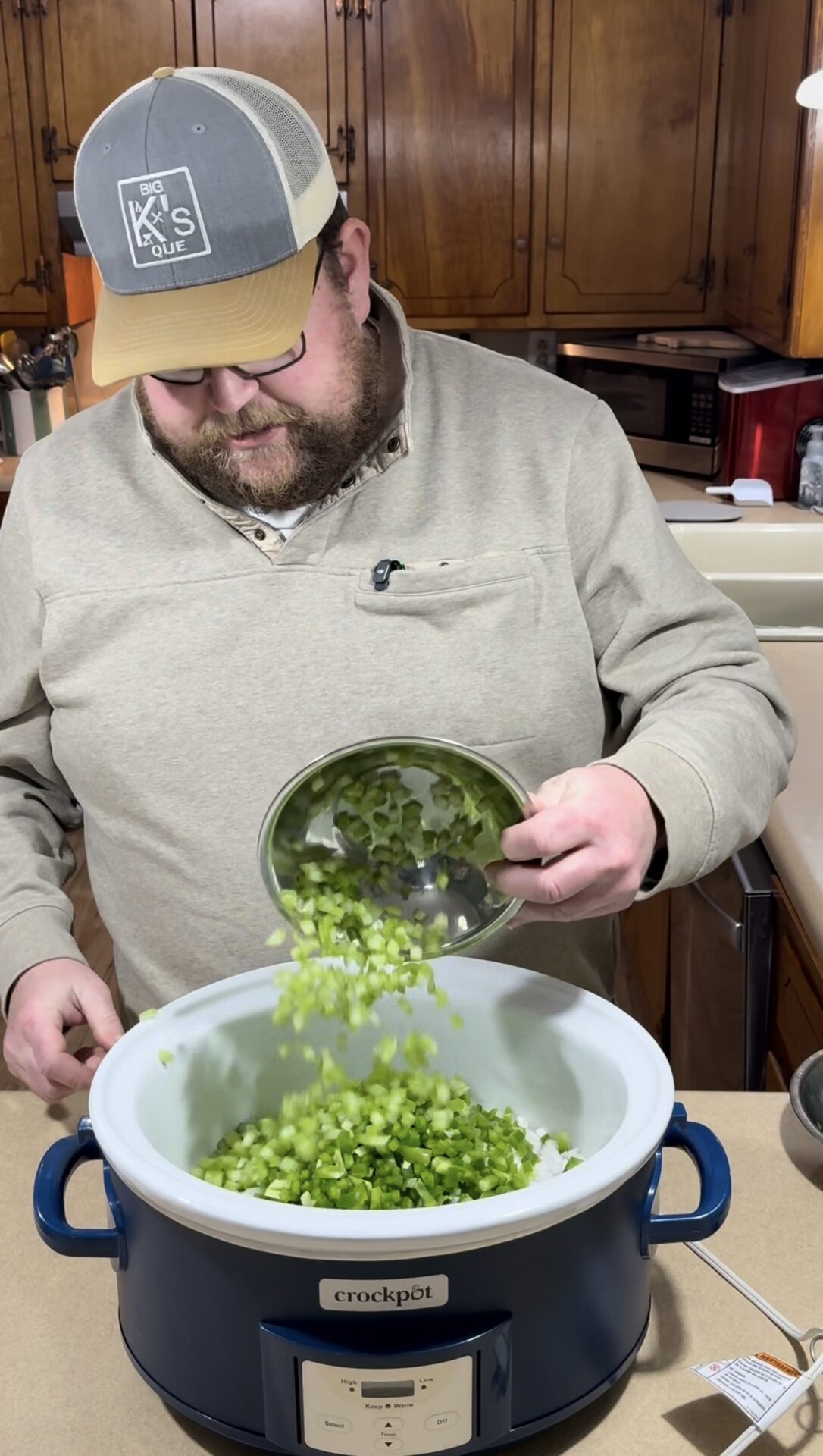 A man wearing a beige sweater and a cap pours chopped celery from a bowl into a large blue Crockpot slow cooker in a kitchen with wooden cabinets.