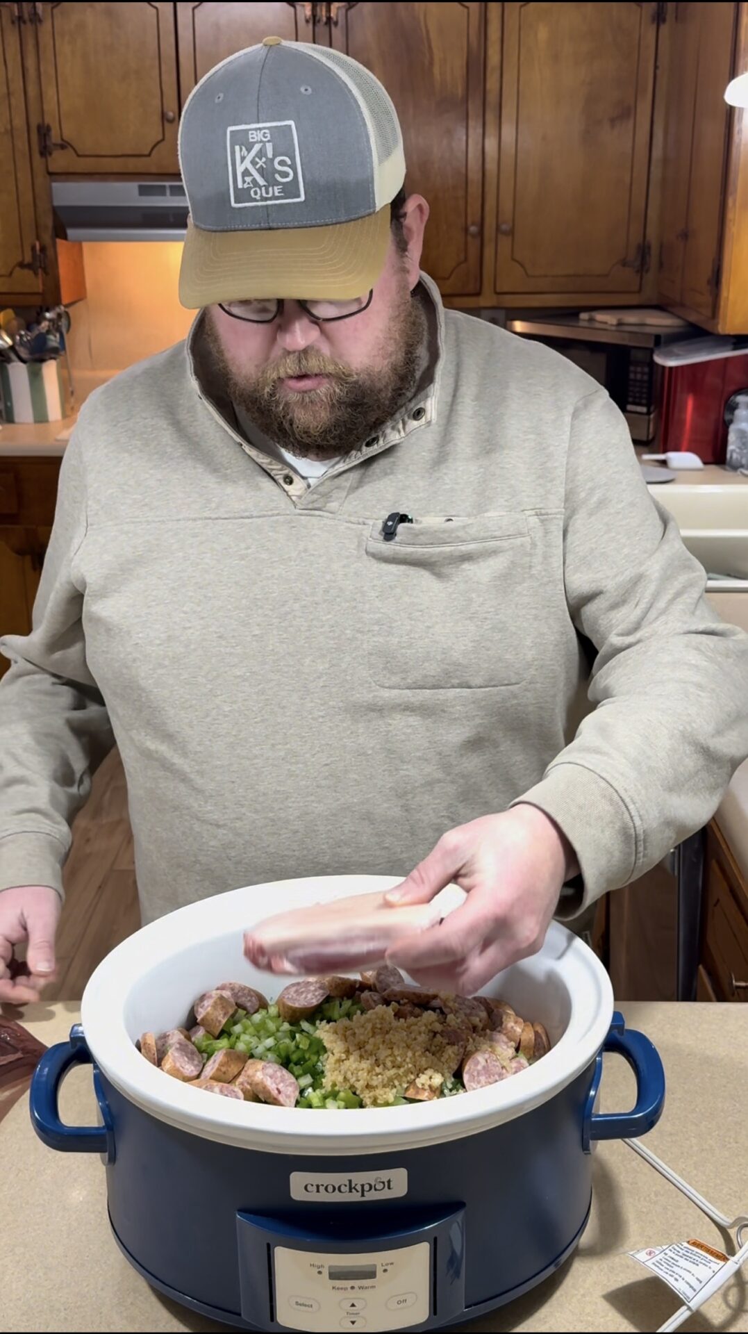 A man wearing glasses, a gray hat, and a beige pullover stands in a kitchen, placing raw pork chops into a crockpot filled with sausage, vegetables, and spices on a counter.