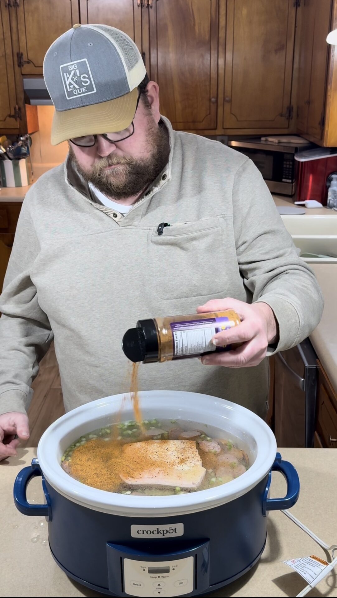 A man wearing glasses and a gray hat sprinkles seasoning from a bottle into a slow cooker filled with meat and broth in a kitchen with wooden cabinets.