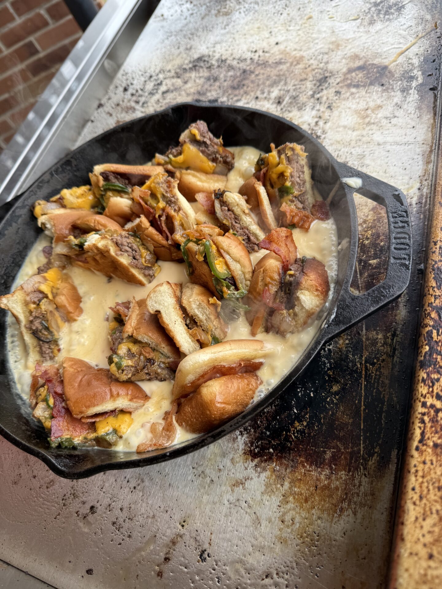 A cast iron skillet on a grill contains chopped cheeseburger pieces with melted cheese and bread, all simmering in a pool of sauce on a greasy surface. Steam rises from the sizzling mixture.