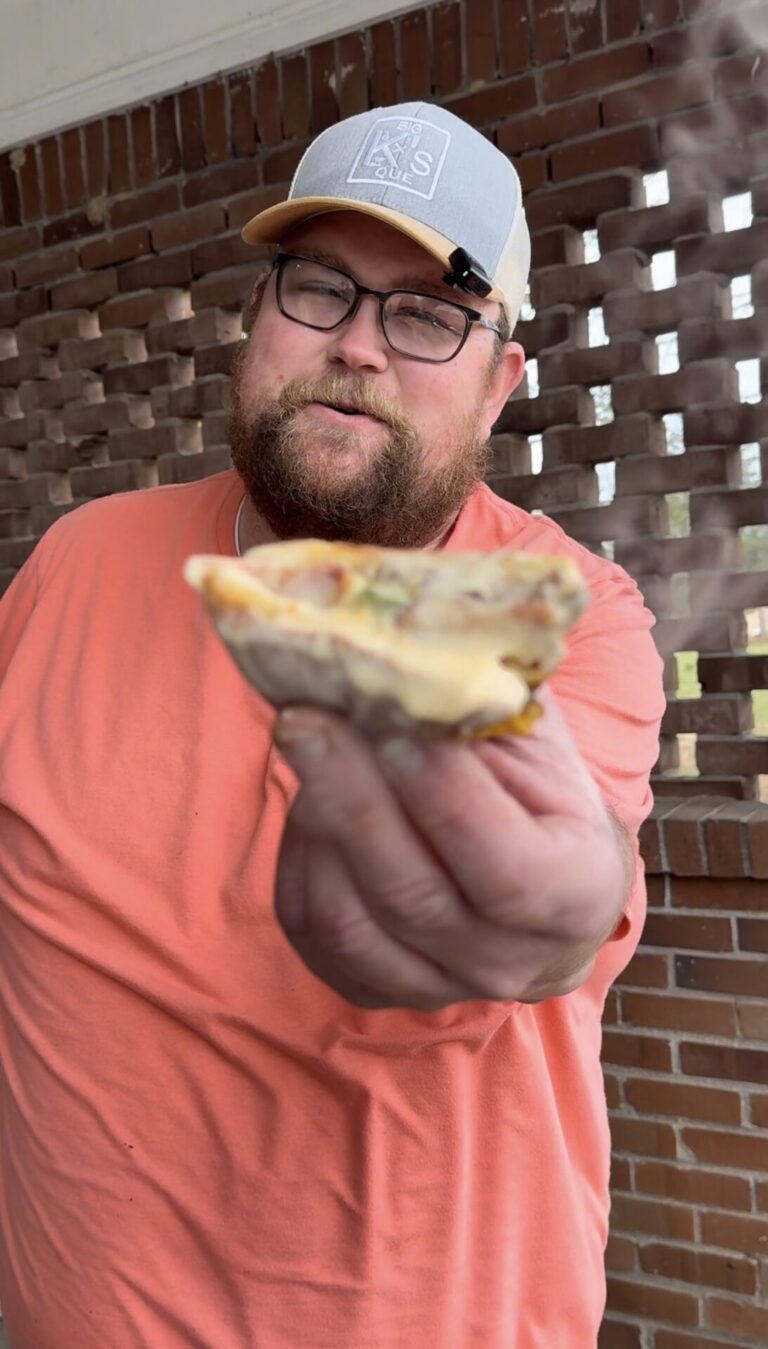 A man in an orange shirt and gray cap holds a steaming slice of pizza toward the camera, standing in front of a brick wall with rectangular openings.