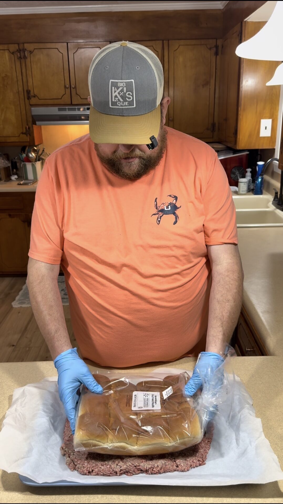 A man in an orange shirt and gray cap stands in a kitchen, wearing blue gloves and holding a large, packaged loaf of bread placed on a slab of raw ground meat on a countertop.