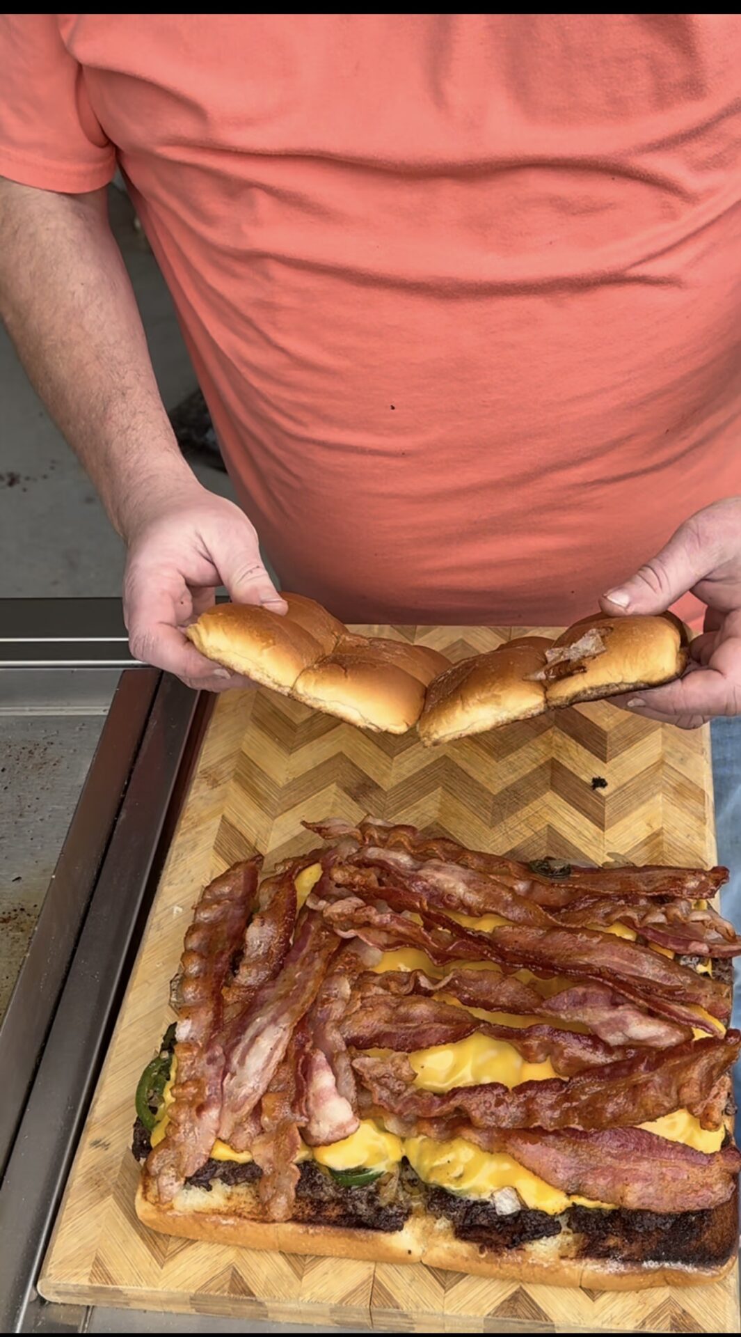 A person in an orange shirt holds a section of sliced sandwich buns above a large rectangular sandwich topped with melted cheese, crispy bacon, and other ingredients on a wooden cutting board.