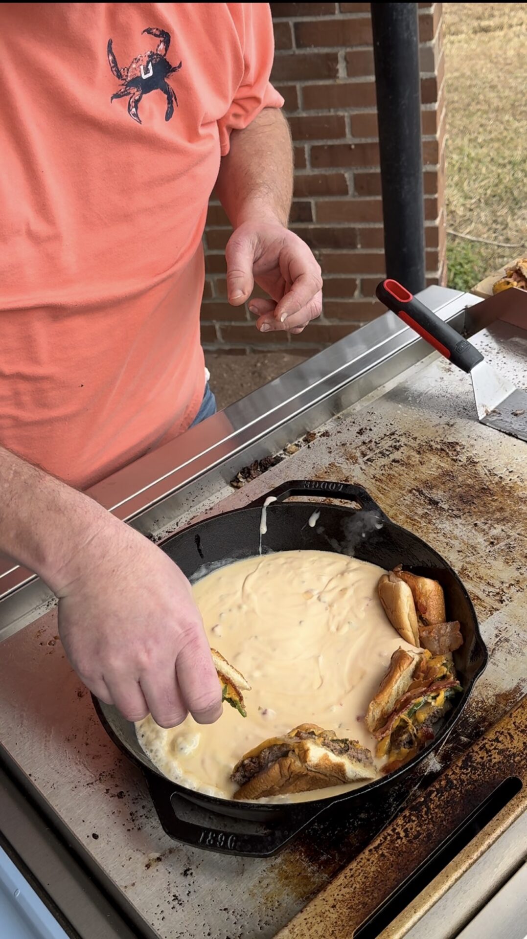 A person in a coral shirt with a blue crab logo dips a sandwich into melted cheese in a cast iron skillet on a griddle. There are several sandwich pieces around the edge of the skillet.