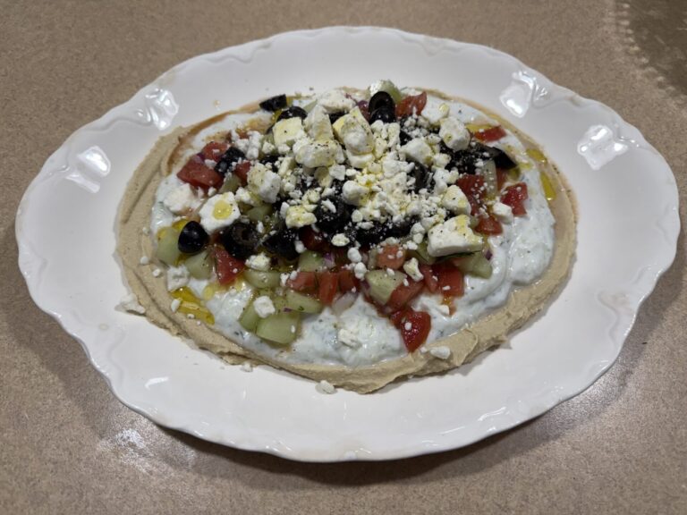 A white plate holds a Mediterranean dip layered with hummus, tzatziki, diced tomatoes, cucumbers, black olives, and crumbled feta cheese, set on a beige countertop.