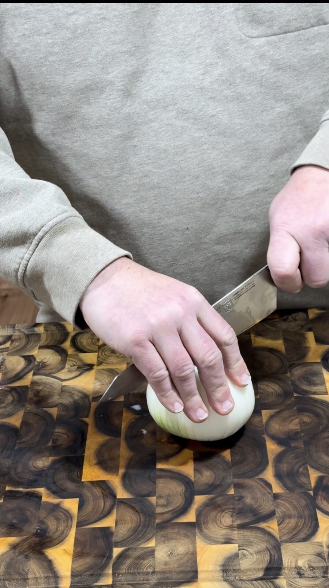 A person wearing a beige sweatshirt slices a white onion with a large knife on a patterned wooden cutting board.