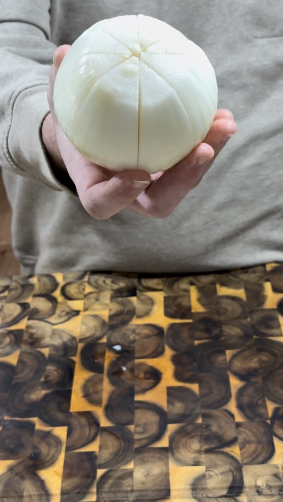 A person holding a peeled onion with crosswise cuts on top, above a patterned wooden cutting board.
