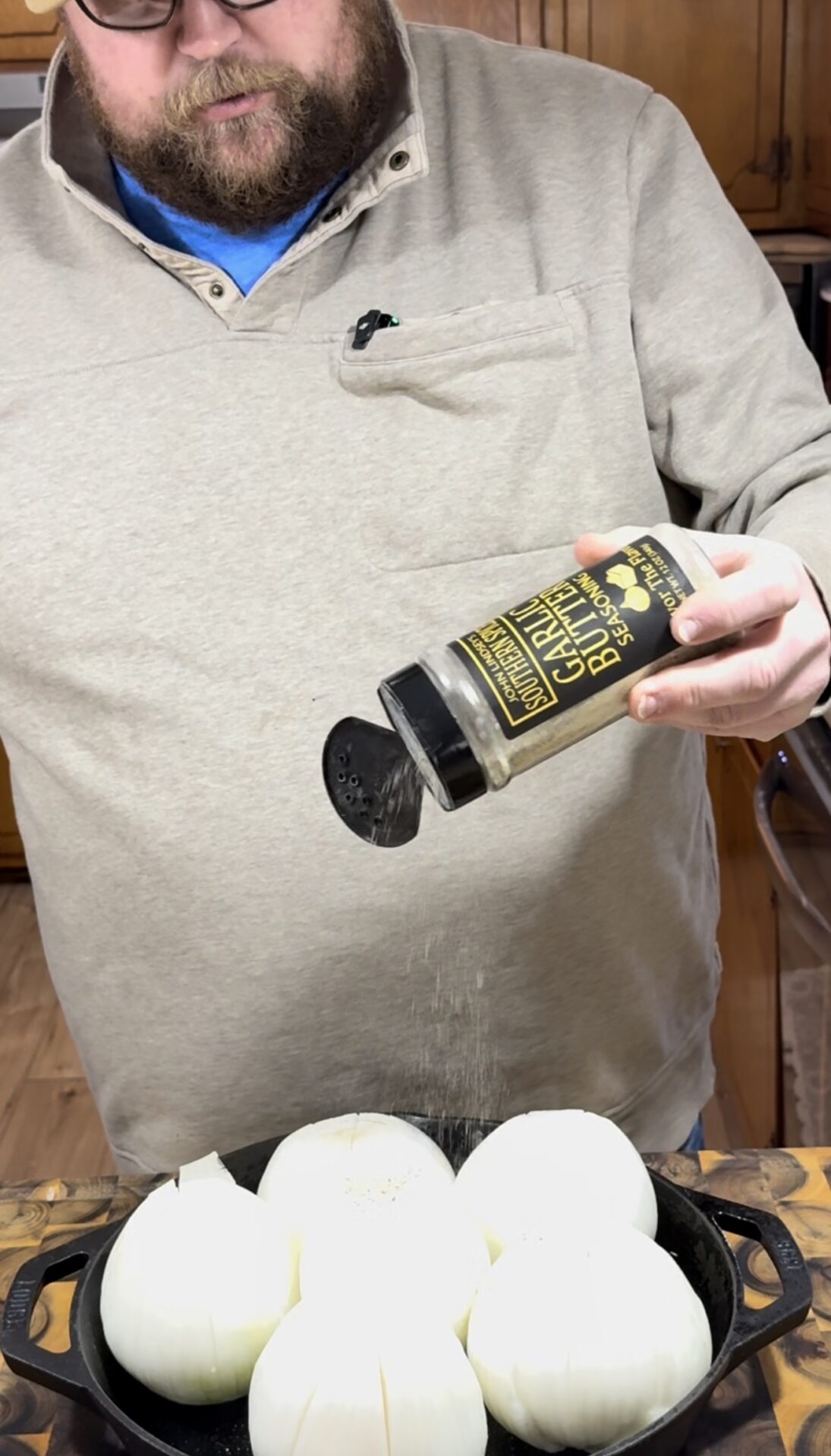 A person sprinkles garlic and herb seasoning from a bottle onto several peeled, cored onions arranged in a cast iron pan on a wooden countertop.