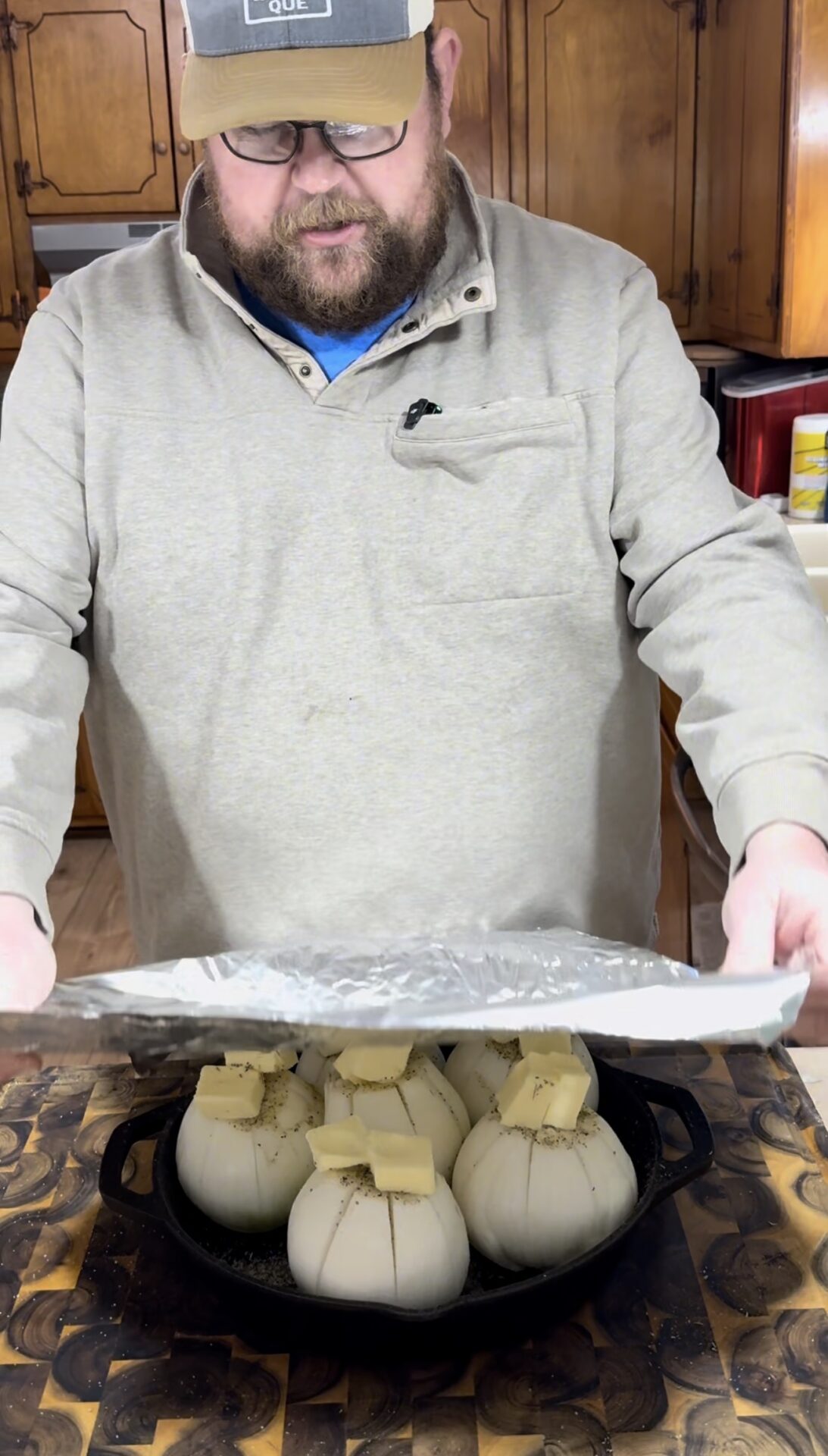 A man in a beige sweater and cap covers a skillet of whole onions topped with butter and seasoning with aluminum foil in a kitchen.